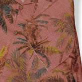 Close-up of the chest area of a Hawaiian shirt, showcasing a reddish fabric with a palm tree pattern in muted browns, greens, and yellows. The detailed print emphasizes the tropical and casual vibe of the garment, with visible button details adding to the shirt&