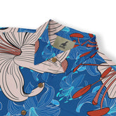 Close-up view of the collar of a Hawaiian shirt, showcasing a vibrant design with beige lilies, red-orange stamen details, and abstract blue floral patterns on a bright blue background, highlighting the "Kalvero" label inside.