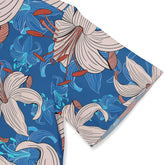 Close-up view of a Hawaiian shirt sleeve featuring a vibrant pattern of beige lilies with red-orange stamen and blue abstract floral designs on a bright blue background.