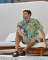 A man sitting on a poolside lounge chair, wearing a matching shirt and shorts set featuring a tropical sea turtle and floral print, exuding a relaxed and cheerful vibe.