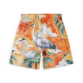 A pair of shorts featuring a vibrant design with cranes standing amidst tropical foliage, including large green leaves and sunflowers. The background is an orange and yellow sunset sky with soft clouds, creating a warm and serene atmosphere.