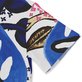 This is a close-up of a shirt sleeve featuring a bold, abstract design with vibrant blue, black, and brown leaves, along with small pink flowers set against a white background. The pattern is dynamic and eye-catching, blending large, striking elements with delicate floral details.