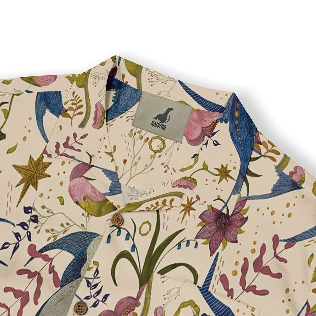 This is a close-up of a shirt featuring a whimsical design with vibrant blue birds and pink flowers, set against a light beige background. The pattern includes various floral elements and stars, with details like wooden buttons and the Kalvero label, enhancing the playful and artistic style of the shirt.